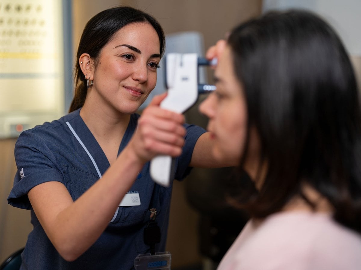 En person i sjukhuskläder undersöker ögat på en patient. Patienten sitter vänd åt sidan, medan vårdpersonalen ler vänligt och koncentrerat genomför undersökningen.