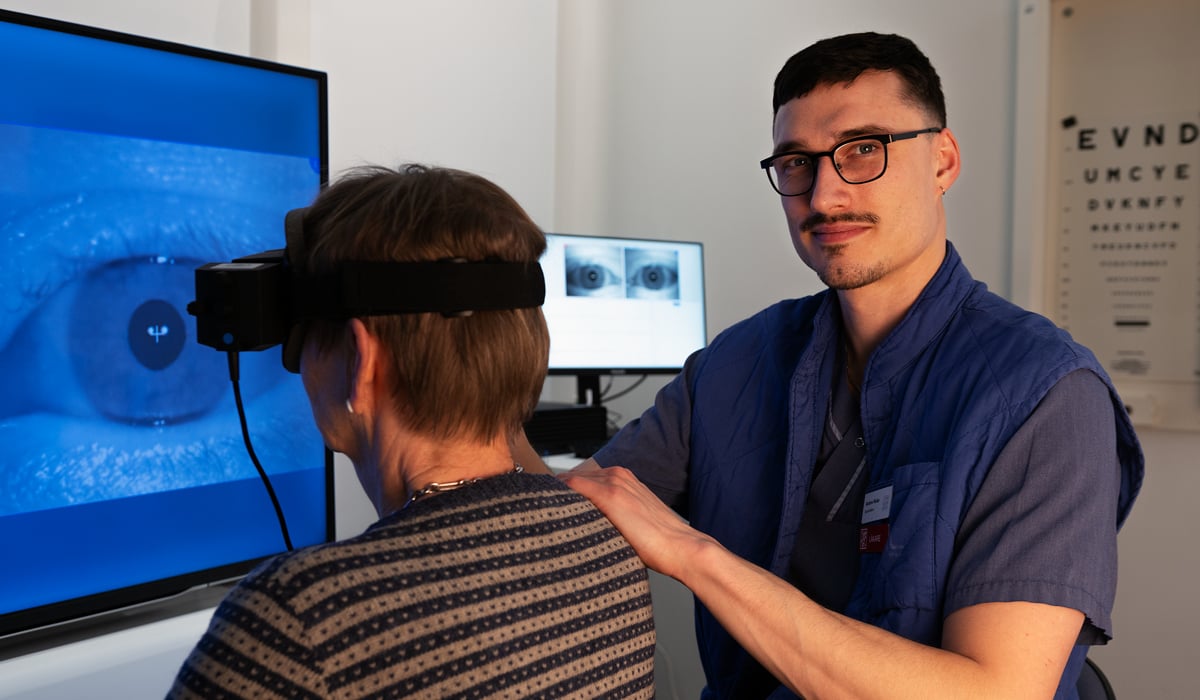 En läkare i vårdkläder bär ett par svarta glasögon. Han håller sin patient på axeln på en patient som har en eye-tracking utrustning på sig.
