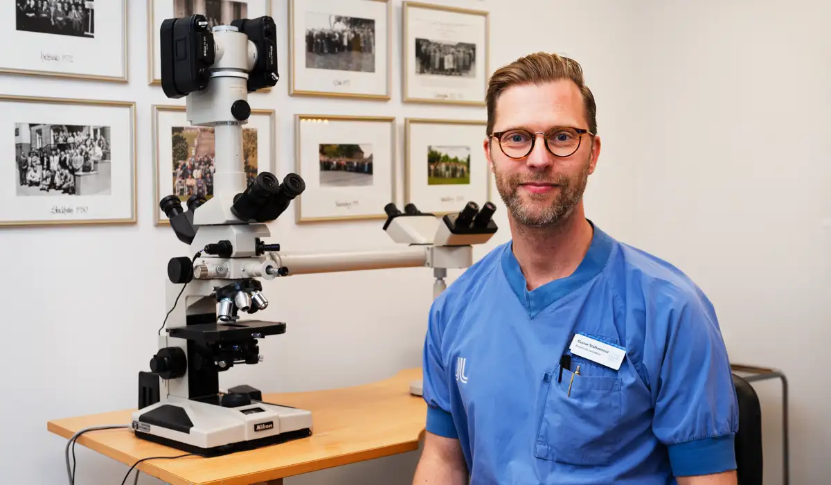 En l&auml;kare i bl&aring; sjukhuskl&auml;der sitter p&aring; en stol framf&ouml;r ett mikroskop.