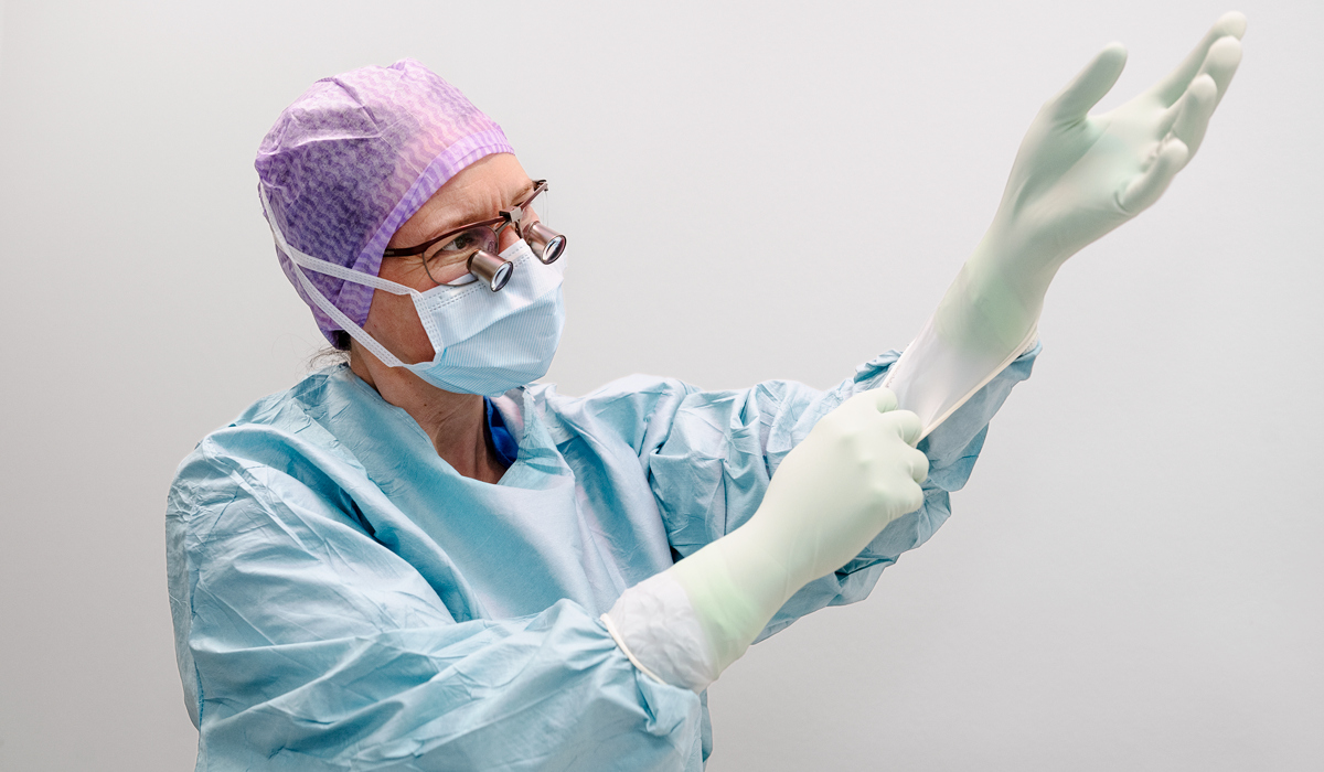 En kvinnlig kirurg b&auml;r operationskl&auml;der, operationsglas&ouml;gon,  munskydd och lila operationsm&ouml;ssa. Hon drar p&aring; sig handskar.
