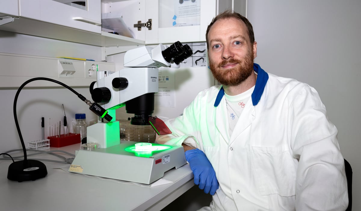 En man i vit labbrock och bl&aring; handskar sitter framf&ouml;r ett mikroskop.