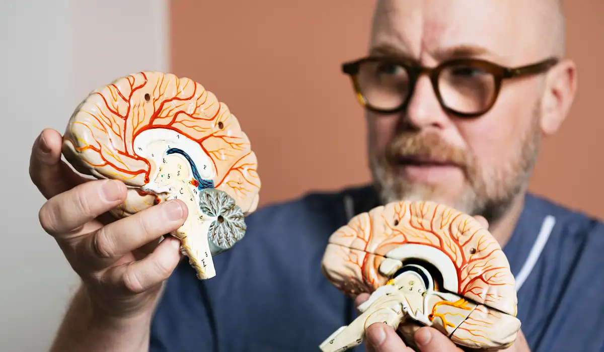 En man i sjukhuskläder och glasögon håller i en modell av en mänsklig hjärna.