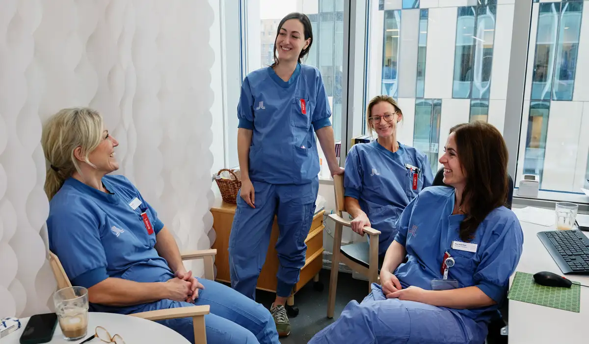 Fyra l&auml;kare i sjukhuskl&auml;der ett rum. Tre kvinnor sitter ner och en st&aring;r upp och h&aring;ller i en stol. De ler mot varandra.