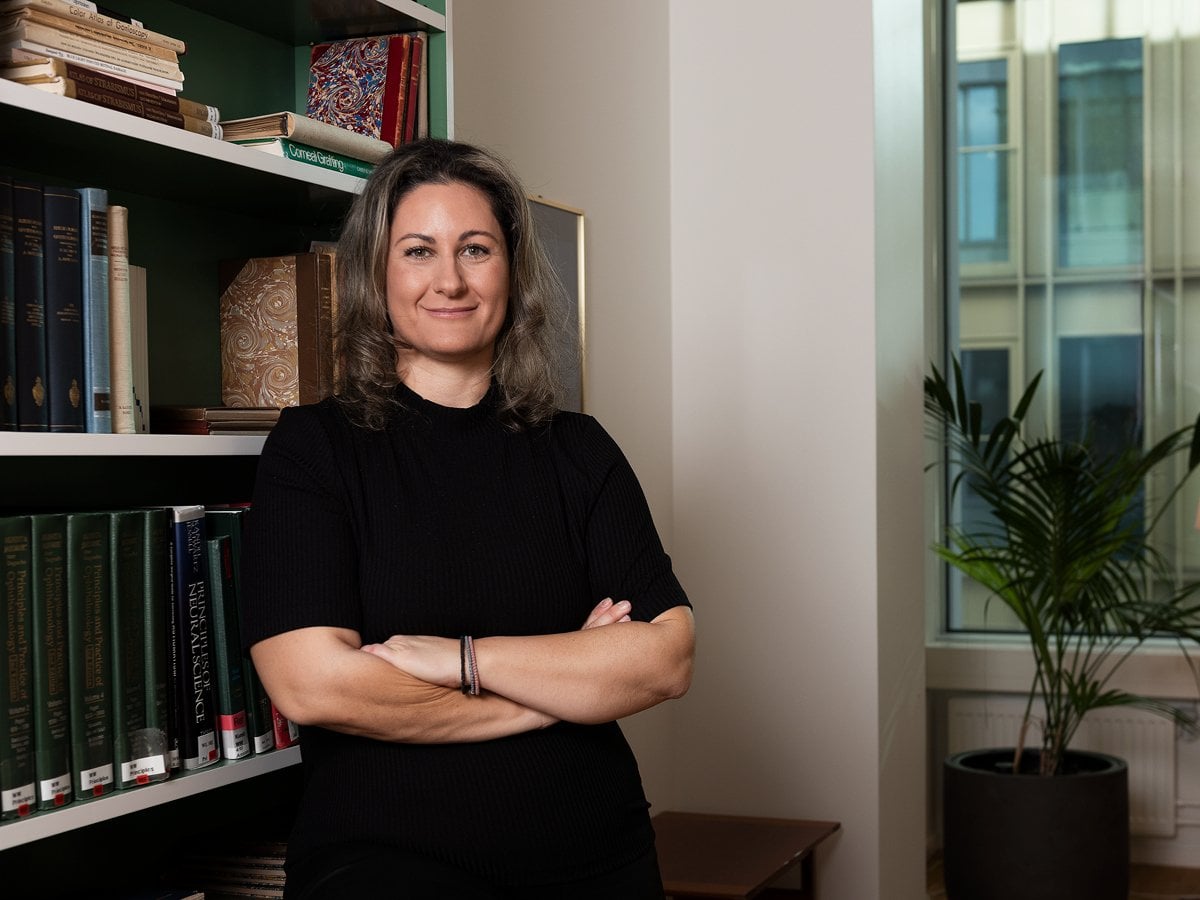 En kvinna i svart klänning håller armarna i kors framför en bokhylla.