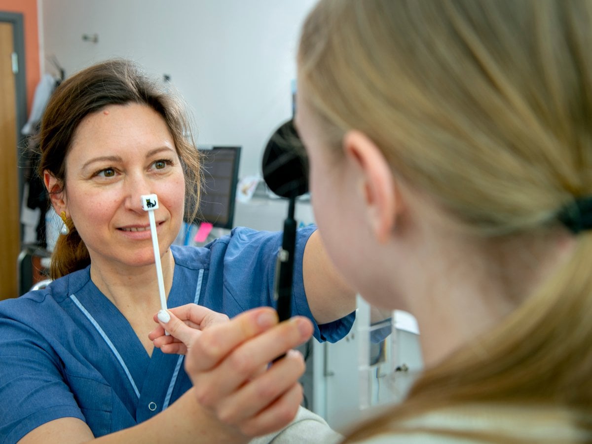 En ortoptist i blå sjukhuskläder genomför en ögonundersökning på ett barn. Hon håller upp  ett instrument framför barnets öga för att undersöka synen.
