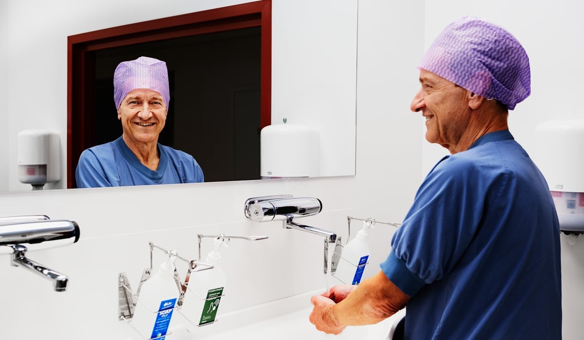 En man står i operationskläder framför en spegel. Han tvättar händerna och tittar i spegeln och ler. 