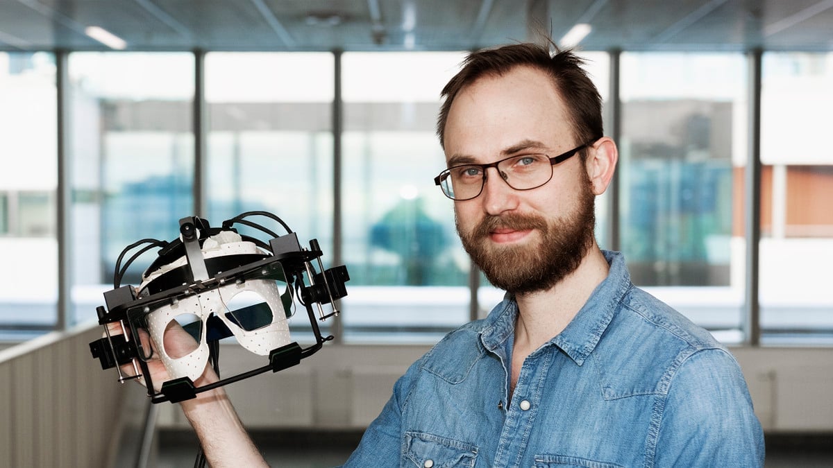 En man bär glasögon, och har en jeansskjorta. Han ler och håller i en ögonrörelse maskin. 