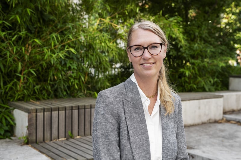 En kvinna med glasögon står framför en träbänk.