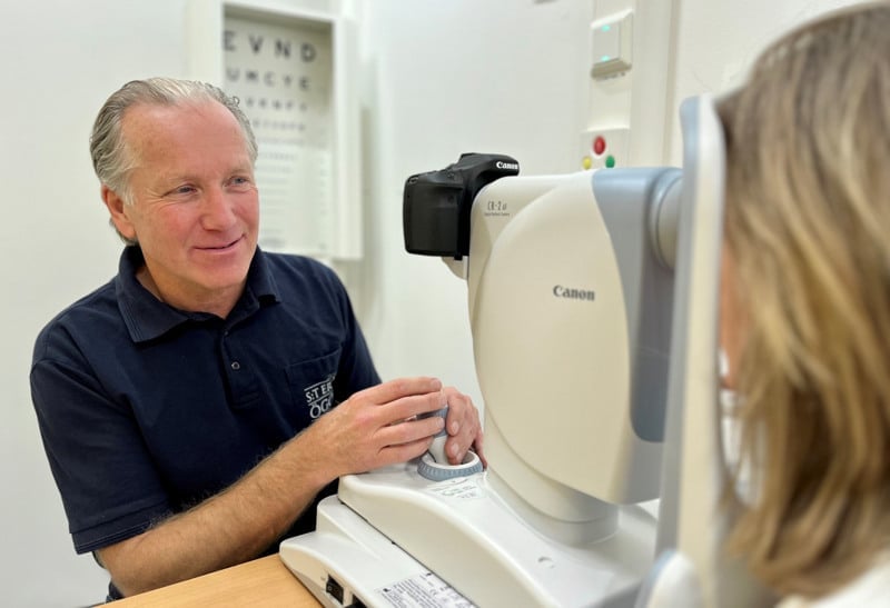 En man i blå pikétröja, sitter vid en  ögonbottenkamera och tittar mot en patient.