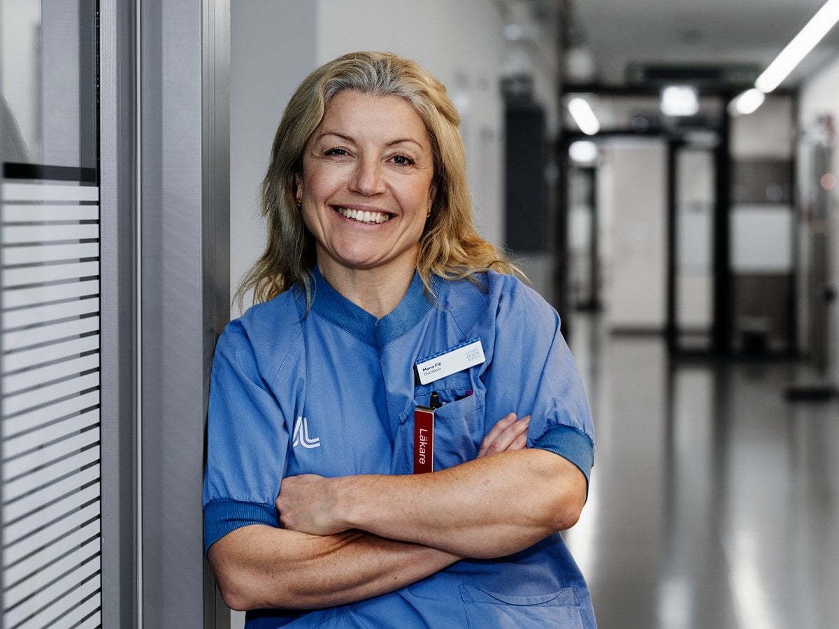 En leende kvinna i operationskläder lutar sig mot en vägg.