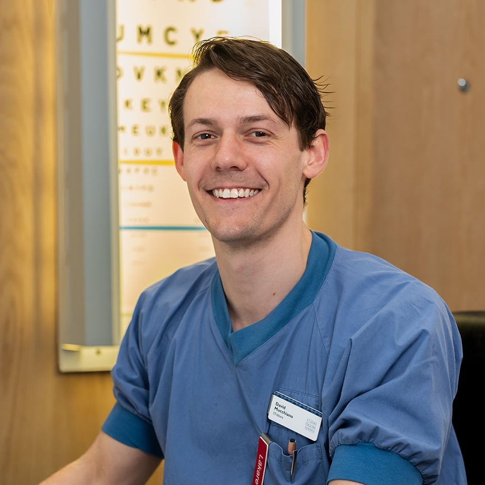 En man i sjukhuskläder sitter framför en dator. Bakom honom syns en syntavla.