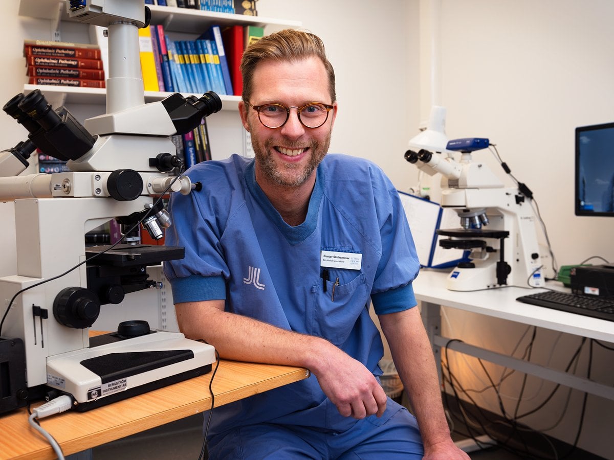 En man i blå sjukhuskläder sitter vid ett mikroskop. Han ler.