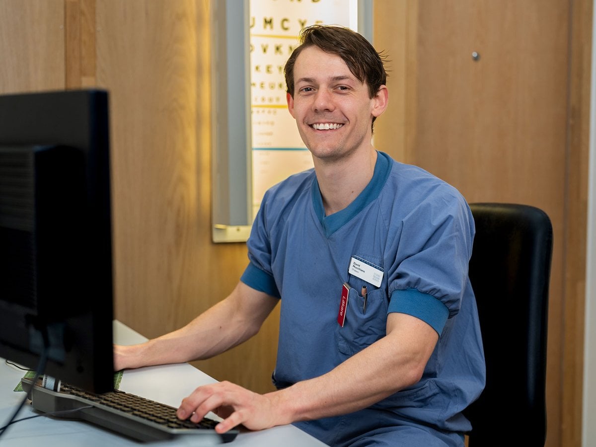 En man i sjukhuskläder sitter framför en dator. Bakom honom syns en syntavla.