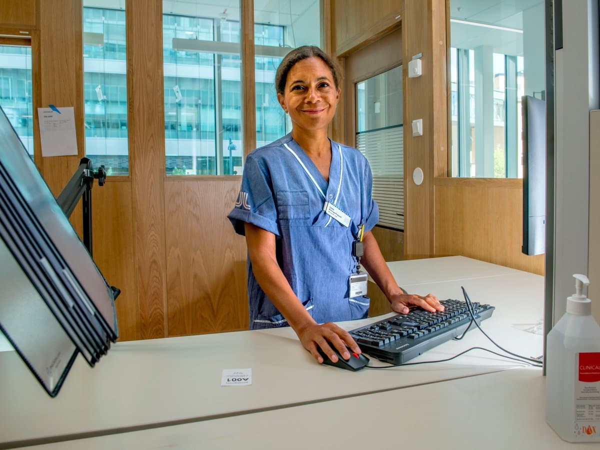 En person i blå sjukhuskläder vid en receptionsdisk. Hennes händer vilar på ett tangentbord. Personen bär en namnbricka och ler mot kameran. Bakgrunden visar en ljus och modern inomhusmiljö med glasfönster och träpaneler.