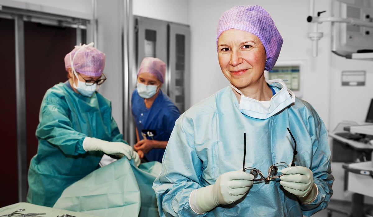 En kvinna i operationskläder håller i ett par operationsglasögon. I bakgrunden syns två medarbetare med operationskläder.