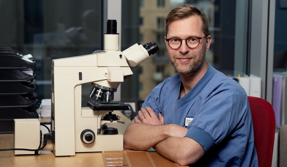 En man med glasögonv och blå sjukhuskläder sitter vid ett skrivbord framför ett mikroskop.