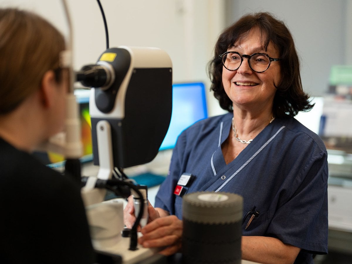 En läkare i sjukhuskläder och glasögon ler medan hon undersöker en patient med hjälp av en ögonlaserapparat. Patienten sitter med huvudet placerat i apparaten för undersökning.