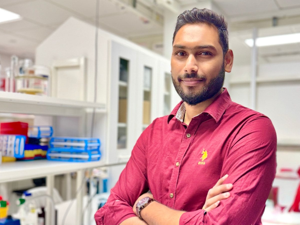 En man iklädd en röd skjorta. Han står med armarna i kors. I bakgrunden syns laboratorieutrustning.