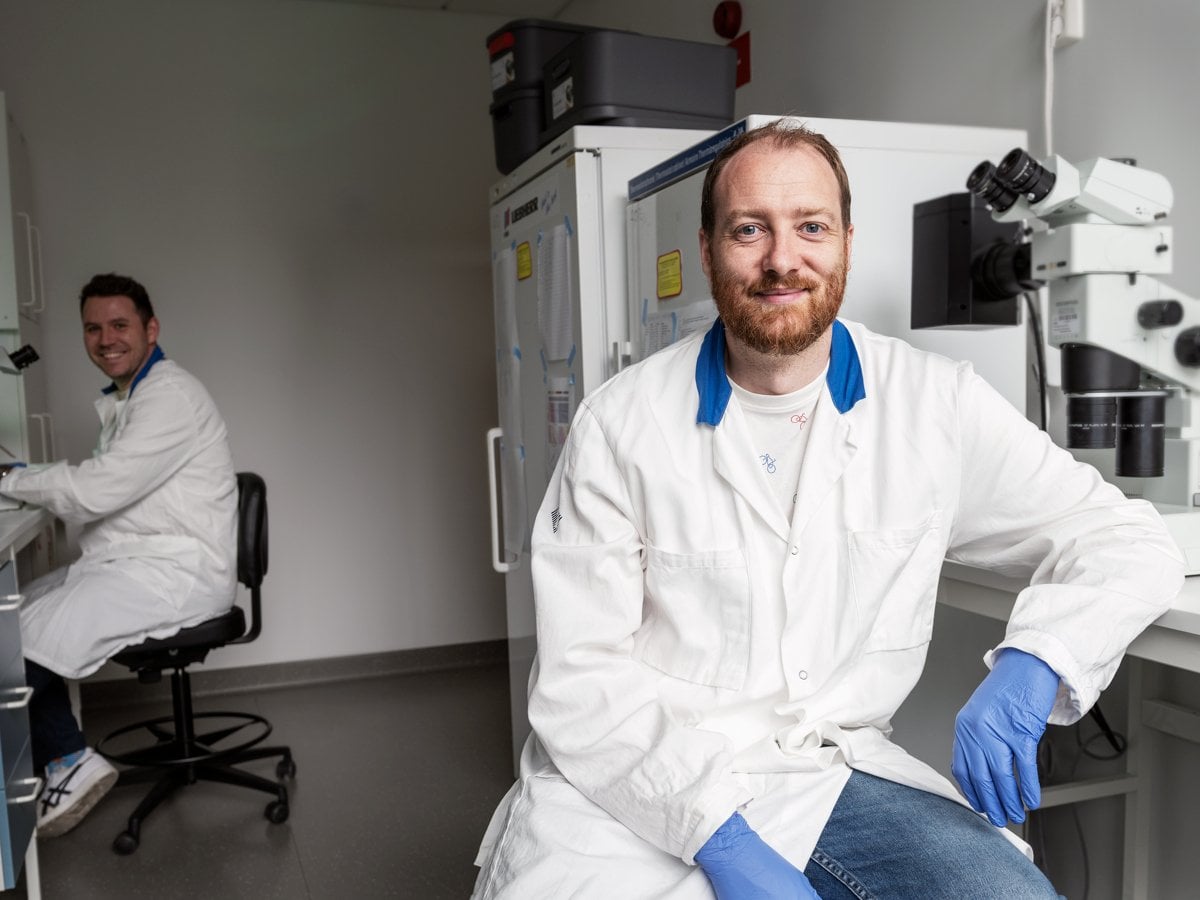 Två män i labbrockar och handskar sitter framför mikroskop.