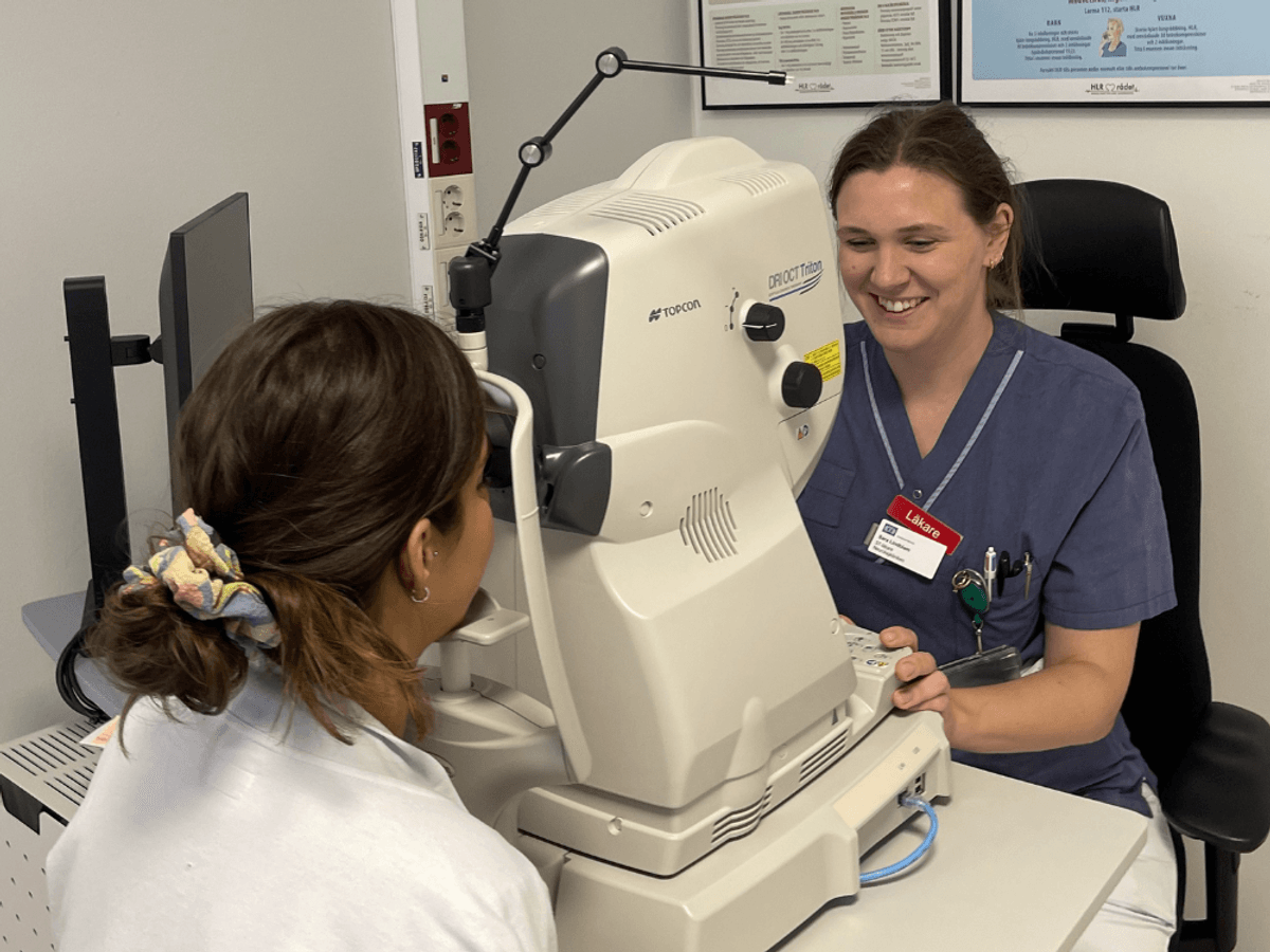 En kvinna i sjukhuskläder ler medan hon utför en ögonundersökning på en patient som sitter framför henne i en ögonundersökningsmaskin.