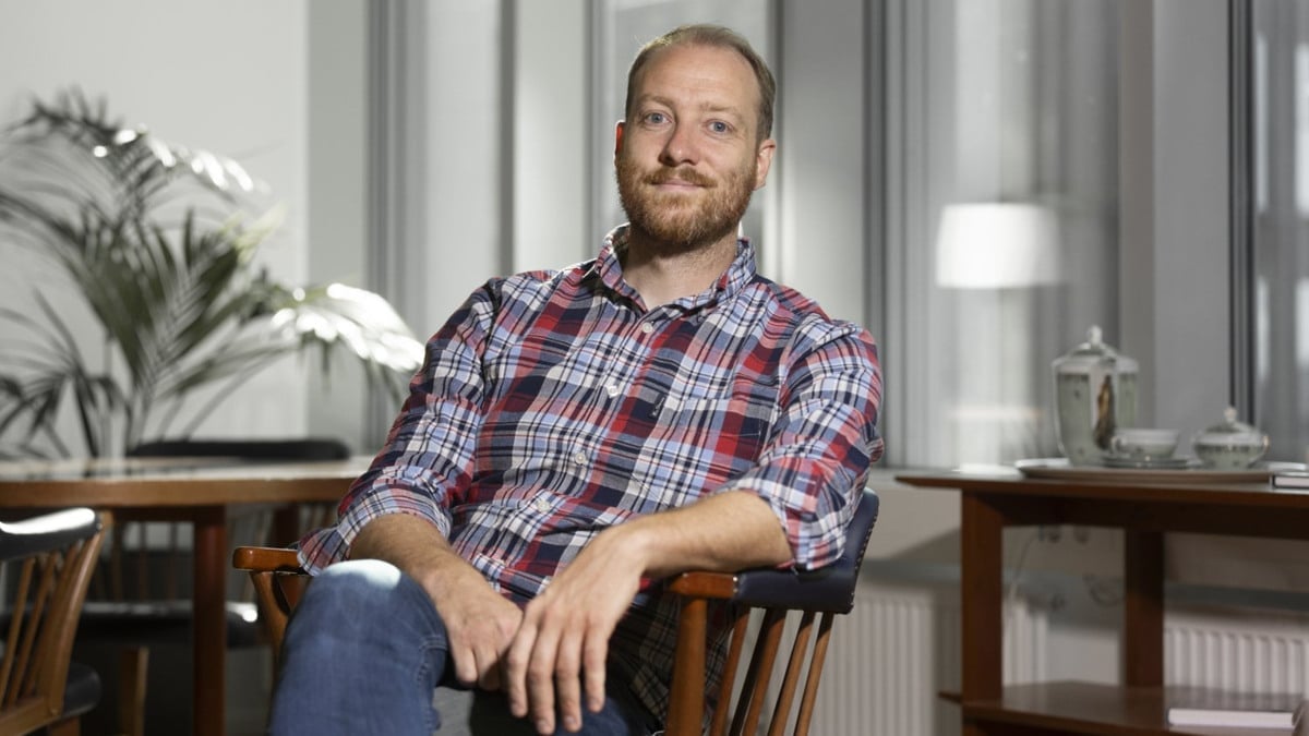 En man som sitter på en stol i ett rum. Han bär en skjorta och jeans.