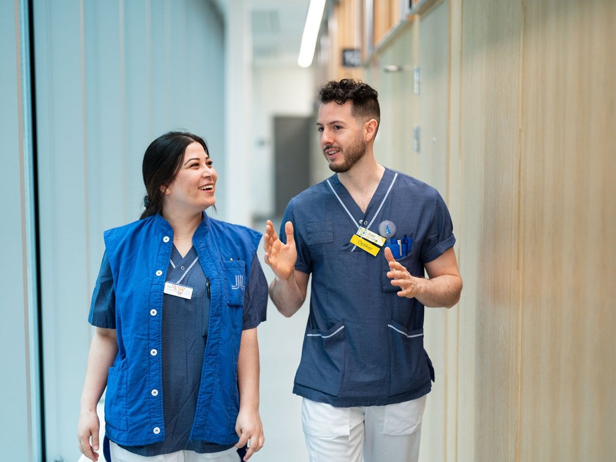 Två personer i blå sjukhuskläder går genom en ljus korridor och pratar med varandra. De ser glada ut och engagerade i samtalet.