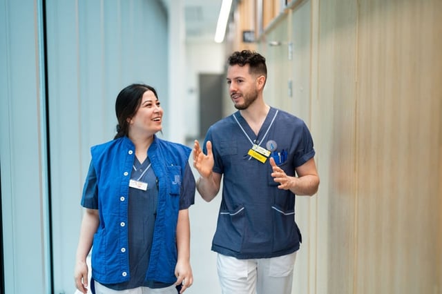 Två personer i blå sjukhuskläder går genom en ljus korridor och pratar med varandra. De ser glada ut och engagerade i samtalet.