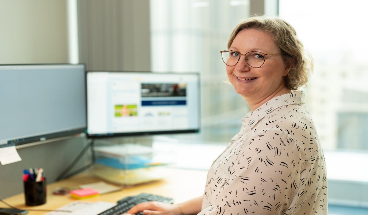 En person med glasögon och ljusmönstrad skjorta sitter vid ett skrivbord med en datorskärm. Hon ler mot kameran.