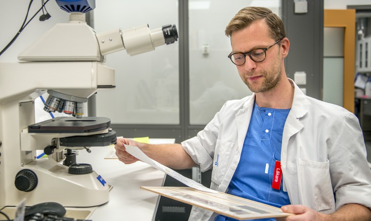 En man i sjukhusuniform studerar en provkarta bredvid ett mikroskop.