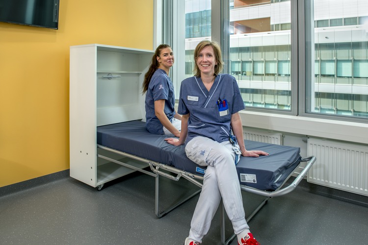 Två kvinnor i blå sjukhusuniform som sitter på en patientsäng och ler.
