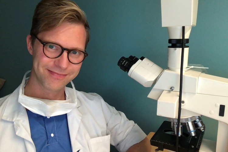 En blond man med glasögon i sjukhusuniform framför ett mikroskop.